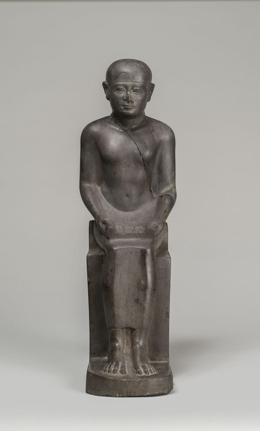 Basalt statuette of Imhotep depicted as a seated figure with a papyrus scroll unrolled across his lap, wearing a tight-fitting kilt and a smooth cap-like wig.
